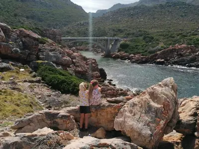 two girls standing on rocks near a river with a bridge at Rocky Bay on the View in Gordonʼs Bay