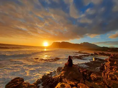 a person sitting on a rocky beach watching the sunset at La Mer Seafront Selfcatering Accommodation Kleinmond in Kleinmond