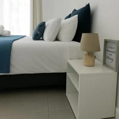 a white bed with pillows and a table with a lamp at Pinelands Apartment in Cape Town
