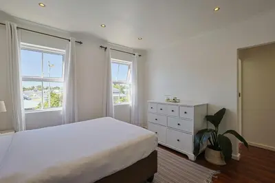 a bedroom with a bed and a dresser and two windows at Fully Equipped Claremont Home with Pool & Style in Cape Town