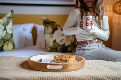 a woman sitting on a bed holding a cup and a plate of food at Paarl Boutique Hotel in Paarl