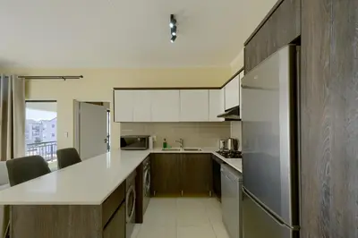 a kitchen with white counter tops and wooden cabinets at Chic & Cozy Apartment with Pool, Gym & Balcony - The Sandown in Cape Town