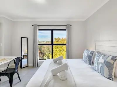 a white bedroom with a bed and a table and a window at Rivers Edge Apartment in Rondebosch with Balcony in Cape Town