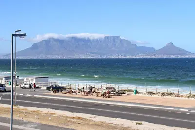 a road next to the ocean with a mountain in the background at Cap Du Mont Cape Town in Bloubergstrand