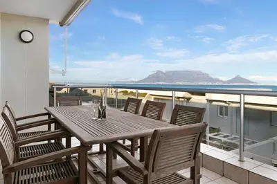 a wooden table and chairs on a balcony with a view of the ocean at Dolphin Beach C111 Sea -Table Mountain Views at Dolphin Beach in Cape Town