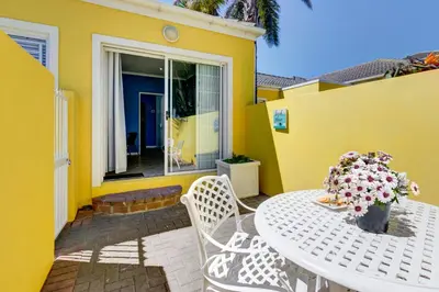 a table and chairs on a patio with a yellow house at Sea Breeze Kite Dreams 1 in Cape Town