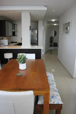 a wooden table with white chairs and a kitchen at Luxurious 3-bedroom Bloubergstrand Cape Town beachfront apartment with stunning sea and beach view in Cape Town
