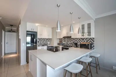 a white kitchen with a large white counter and stools at Modern 2 Bedroom Apartment At Dolphin Beach in Cape Town