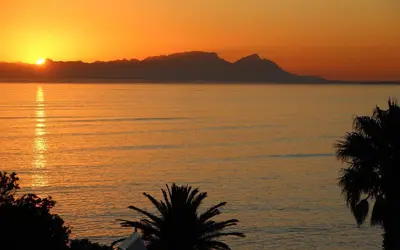 a sunset over a body of water with palm trees at 'Die Braaihuis'- sleeps 6, Large Veranda, Sea Views, Central and Fabulous! in Gordonʼs Bay