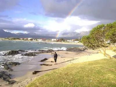 a man fishing on the beach with a rainbow in the background at 'The View' on Bikini Beach, Seafront, 2 bedroom, Sleeps 4 in Gordonʼs Bay