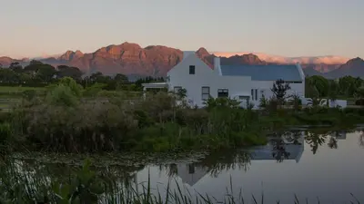 a white house with mountains in the background next to a lake at Sanddrif Stellenbosch Guest Farm in Stellenbosch