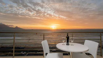 a table with two wine glasses on a balcony with the sunset at The Waves Blouberg Beach Apartments in Bloubergstrand