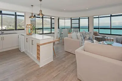 a kitchen and living room with white furniture and windows at The Sun, Whales & Waves - Seafront Apartment in Hermanus