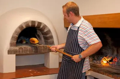 a man in an apron putting something into an oven at Penguino Guesthouse in Hermanus