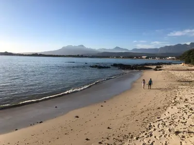 two people walking on a beach next to the water at By Die See in Gordonʼs Bay