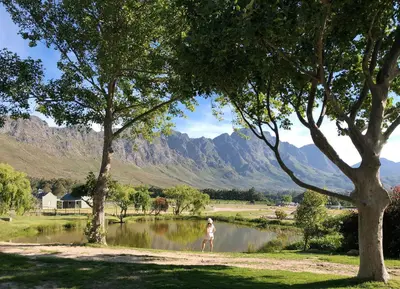 a woman walking by a lake with mountains in the background at Jardin de la Ferme in Franschhoek